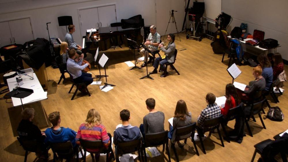 Five musicians perform various stringed and horn instruments to an audience of young people as part of Bristol music charity, Sound World. There is a row of about 11 people watching the performance, which is taking place in a hall.