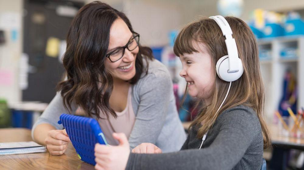 Teacher and child in a classroom. The child is wearing headphones and holding an ipad. They are both smiling.