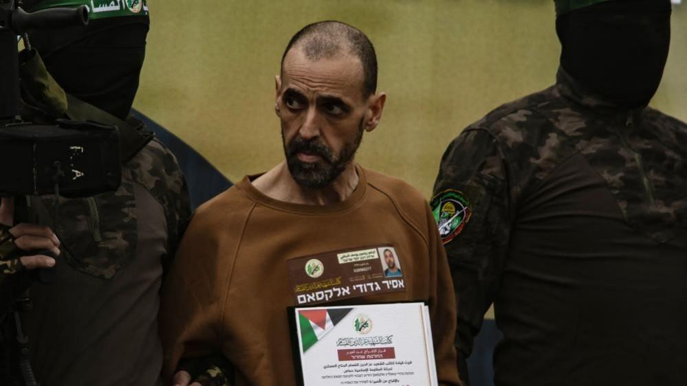 Sharabi, wearing a brown sweatshirt adorned with identification, stands between two masked Hamas men.