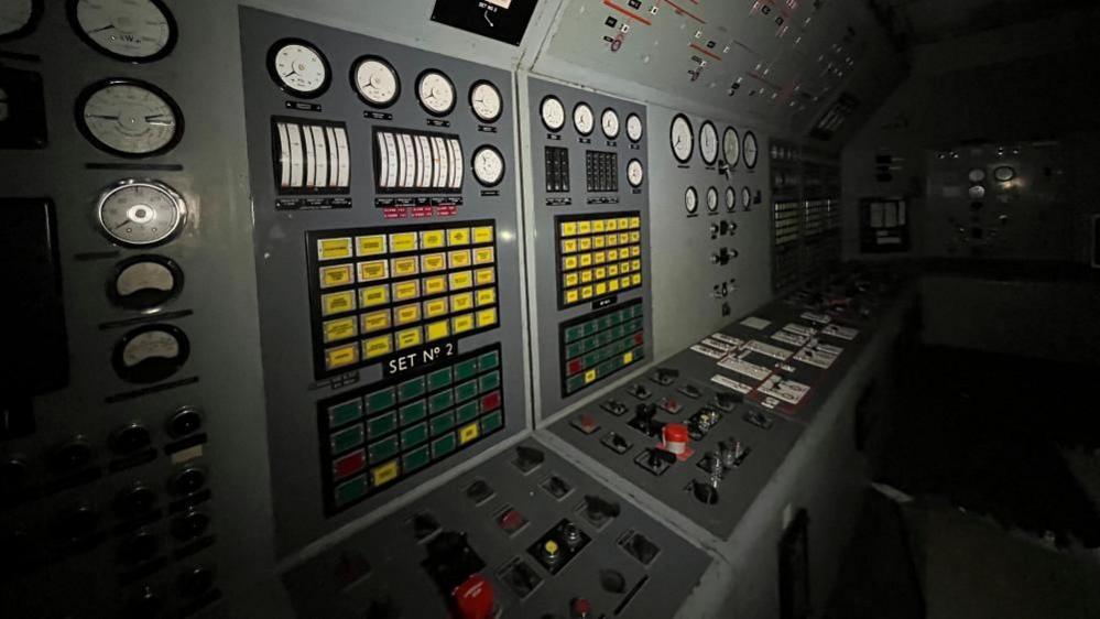 A control panel of switchgear inside a power plant control room. Many of the buttons are coloured and they are housed onto grey cabinetry.