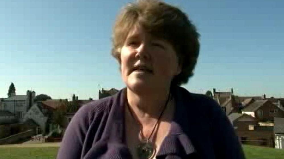 Rebecca Breese with fair hair wearing a purple top and a necklace with a large pendant, standing in a grass park with buildings behind