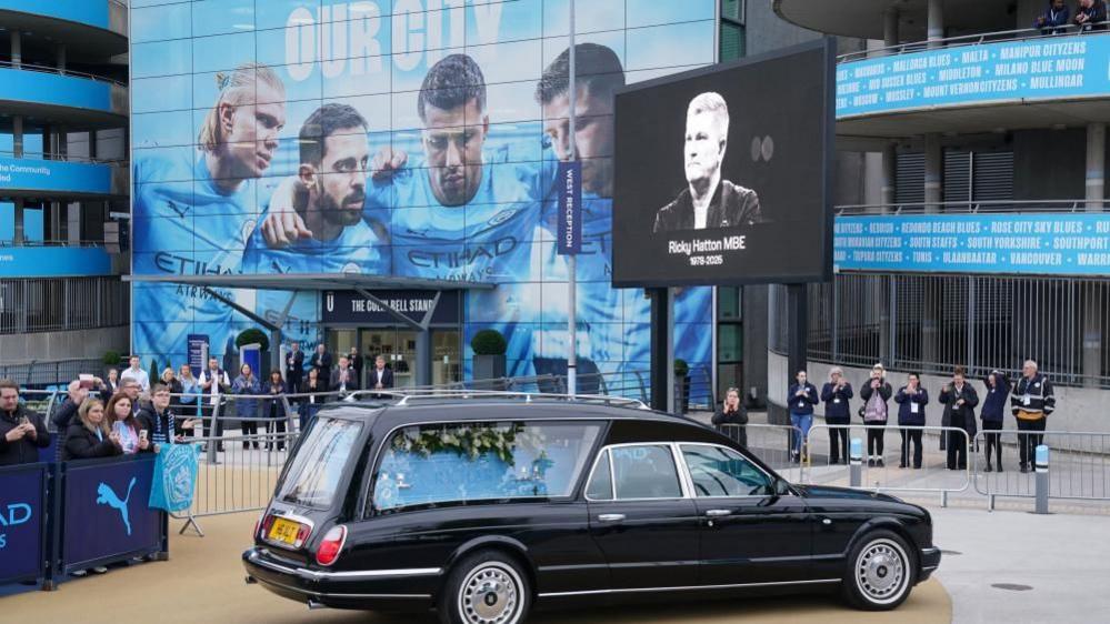 A black hearse with a blue coffin covered in flowers is outside a football stadium. People stand nearby behind barricades.
