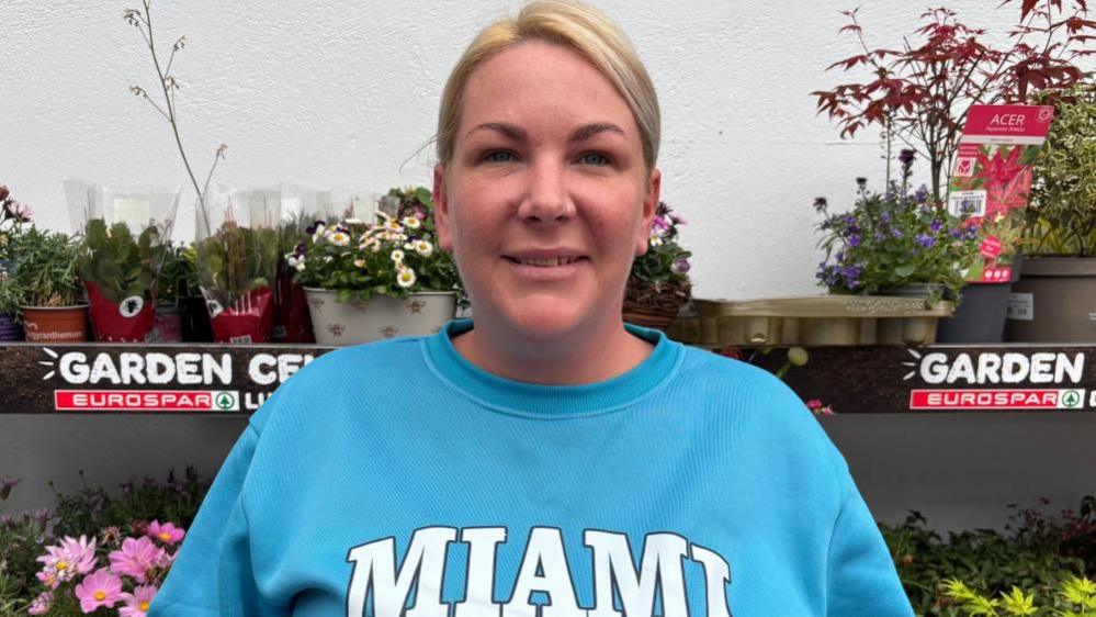 A woman with blonde tied back hair smiles into the camera. She has a nose ring and is wearing a light blue sweatshirt that says 'Miami' in capital letters. She is standing in front of a stall of flowers in plastic pots.