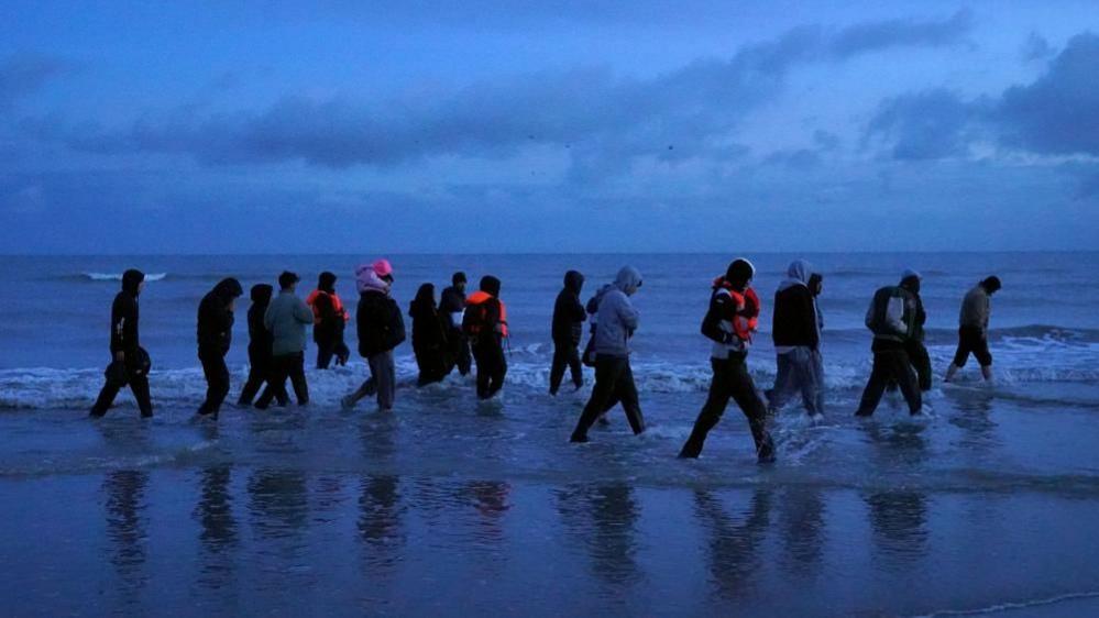 A large group of migrants are seen wading through the shore to board a small boat in France