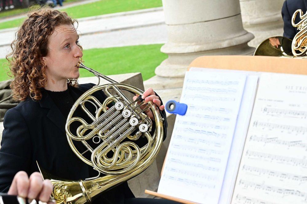 A person with long brow hair is playing a French horn. 
A sheet of music is visible beside them.