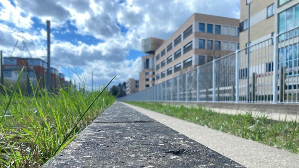 A strip of grey concrete bordered by green grass. Building line either side of the strip. On the right-hand side in a grey metal fence. In the distance is a cloudy sky.