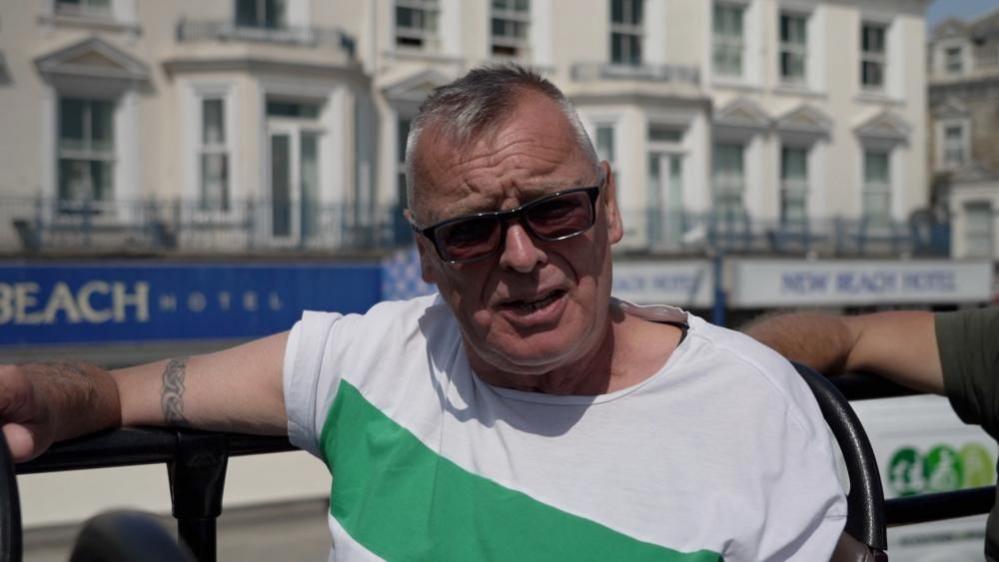 Michael Wilson is sitting on an open top bus, wearing sunglasses. He has short grey hair, is clean shaven, and is wearing a white and green striped top. The bus is moving past the New Beach Hotel, signage for which can be seen in the image.