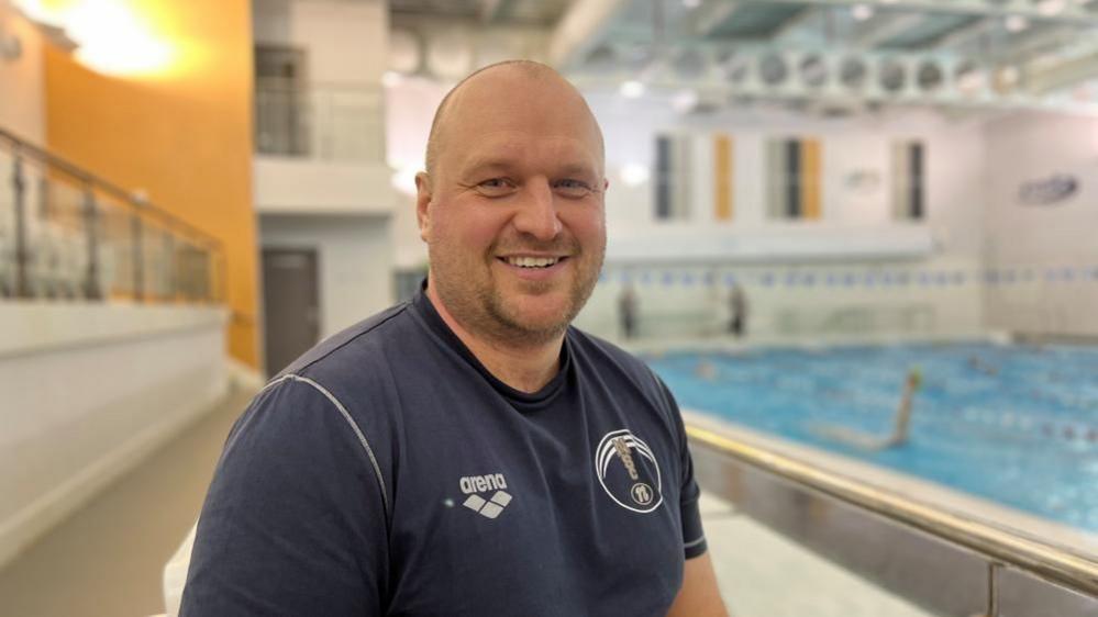 Andy Sharp with very short hair and slight beard wearing a blue T-shirt and sitting in a white chair alongside a swimming pool. A swimmer doing back stroke is visible behind him.