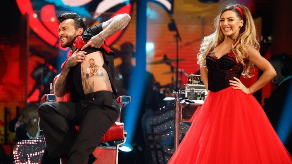 Pete Wicks with medium-length swept-back black hair, wearing a black shirt, red scarf and black trousers, performing a twisting manoeuvre with Jowita Przystal with long blonde hair wearing a dress with black top and red skirt. The Strictly band is visible behind them with art deco shapes on the scenery.