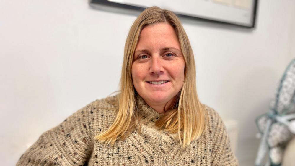  Emma Spencer is sitting at a table at the Lakenheath Community Center wearing a thick, beige knitted sweater with a speckled pattern. In the background, part of a framed picture is visible on a white wall. Emma is smiling at the camera and look relaxed.