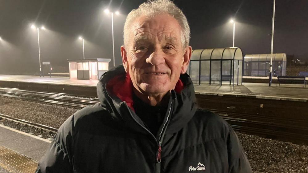 A man in his 70s wearing a coat standing on a railway station platform.