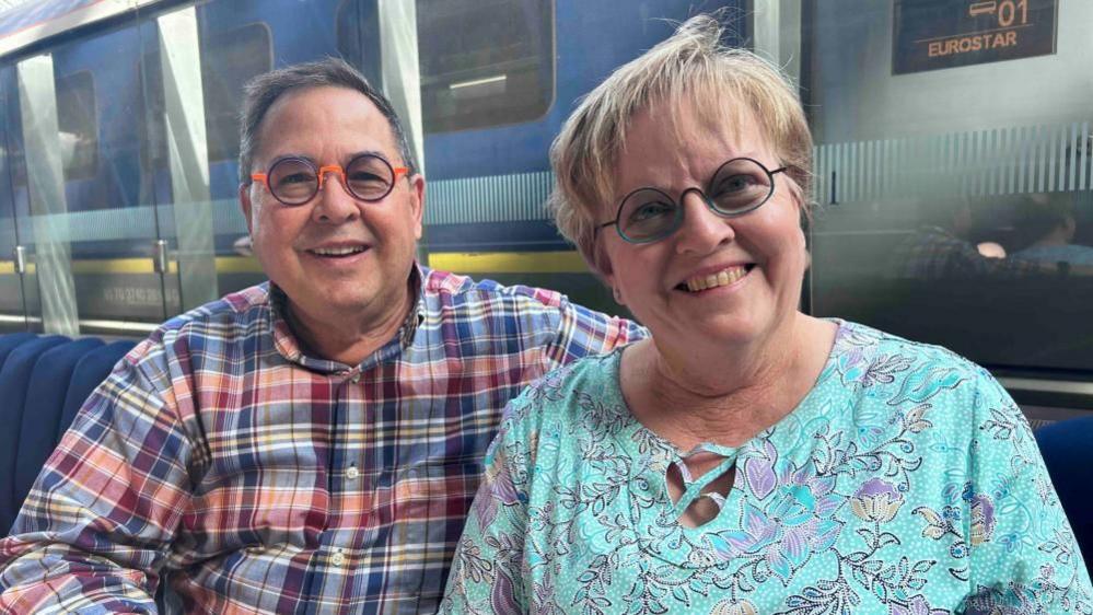 Rand and Janeel Smith, at St Pancras railway station, next to a train, smiling at the camera. Rand is wearing a red and blue checked shirt, and glasses and Janeel, a blue top with flowers on, with shirt fair hair and glasses. 