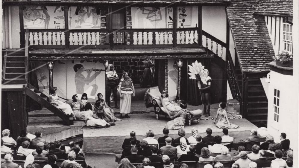 A black and white photograph showing actors on an outdoor stage performing to an audience at the George Hotel. 