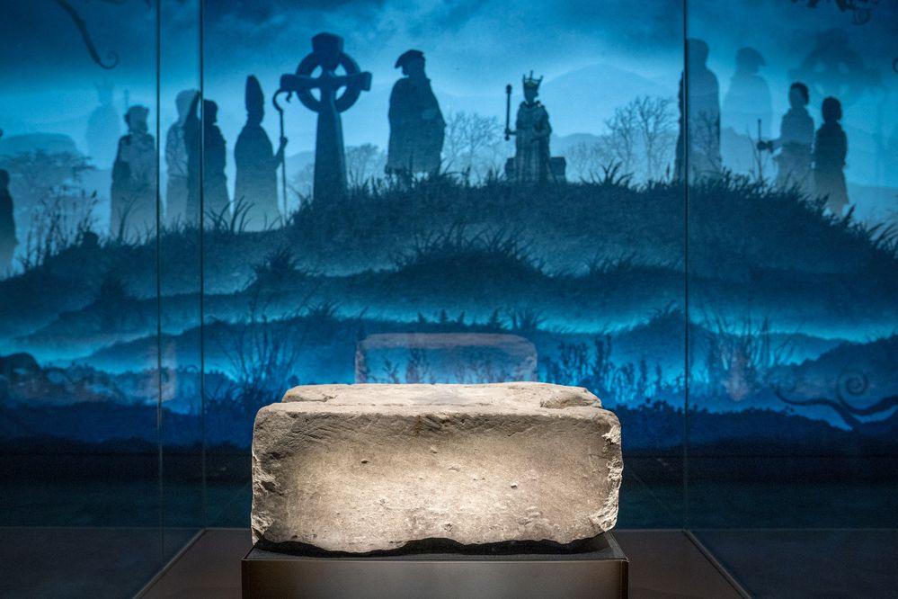 The Stone of Destiny on display at the new Perth Museum. A blue-shaded backdrop behind the stone shows an illustration of old Scottish royalty gathered on a hill. 