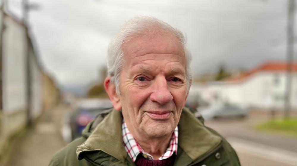 Gerald Kelly is standing on a paved street with buildings and parked cars in the background. He is wearing a green jacket over a red-and-white checked shirt. The sky appears overcast.