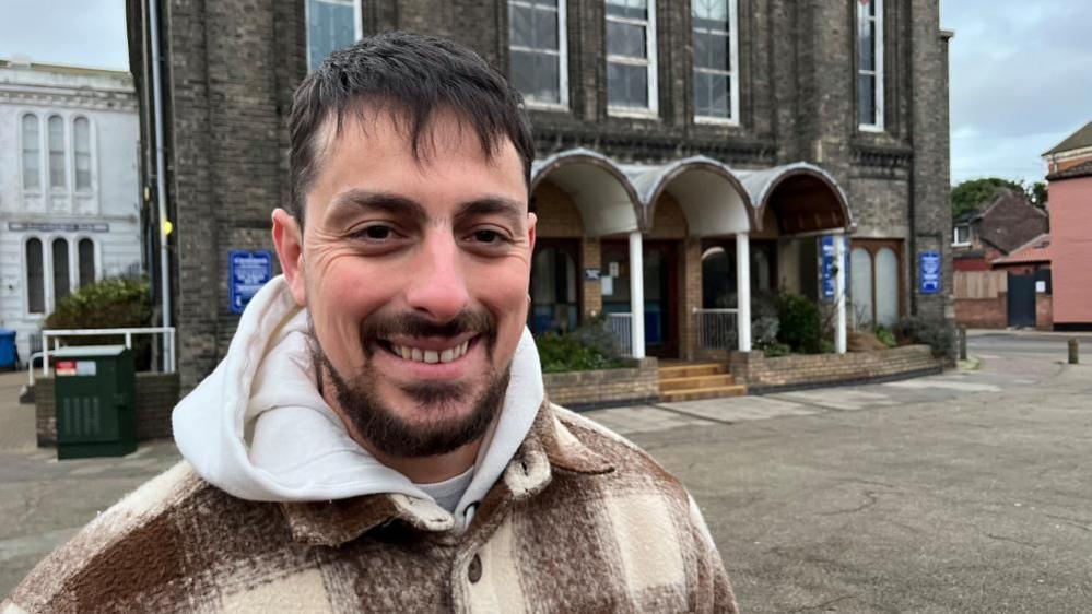 Ruben Cruz, who has a beard, moustache and dark hair, wearing a cream hooded top and brown and cream chequered jacket. He is standing outside the Christchurch building in Great Yarmouth.