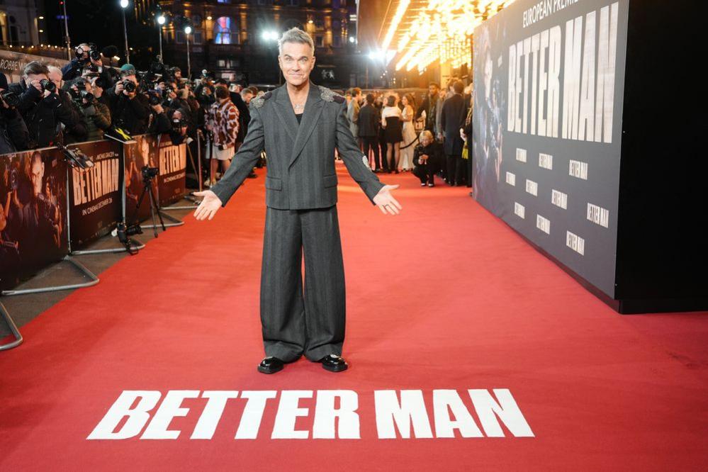 Robbie Williams wears a dark striped, slightly baggy suit on the red carpet for his film Better Man. Photographers can be seen behind the barrier in the background. Robbie gestures towards the red carpet in front of him which has the words "Better Man" written on it in white letters. 