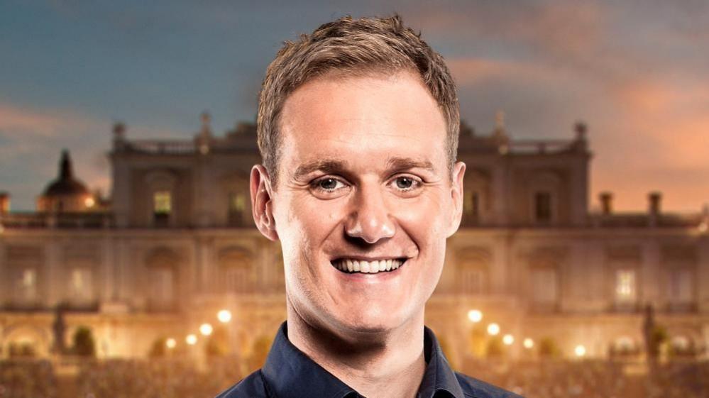 A man stands in front of a palace. He has short blonde hair and is smiling at the camera.