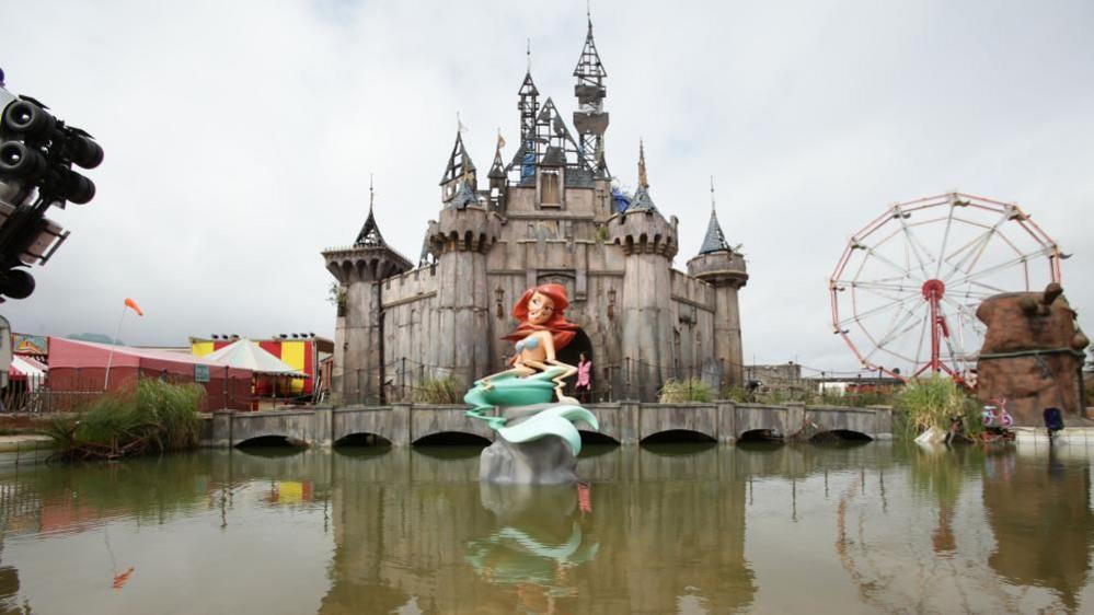 Banky's Dismaland bemusement park, which features a grey Disney-style castle, Ariel the mermaid and a ferris wheel in front of a lake.