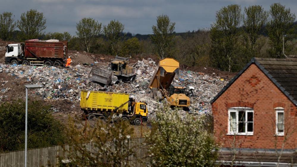 Three trucks emptying tonnes of rubbish at a landfill site on a hillside behind a house. There is a plough in the mound of rubbish alongside them.