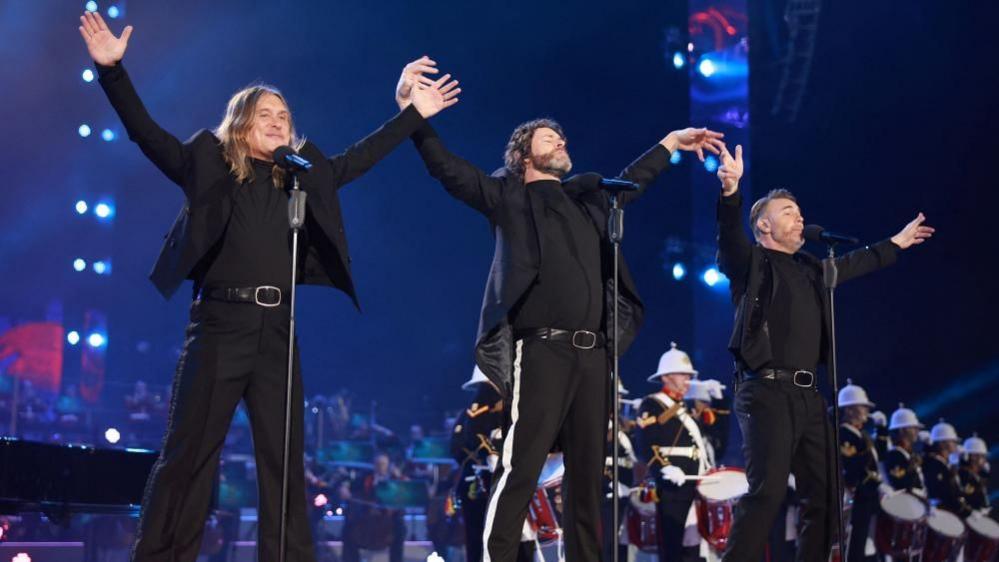 Mark Owen, Howard Donald and Gary Barlow from Take That perform on a stage. They each stand in front of microphones on stands and have their arms outstretched. 