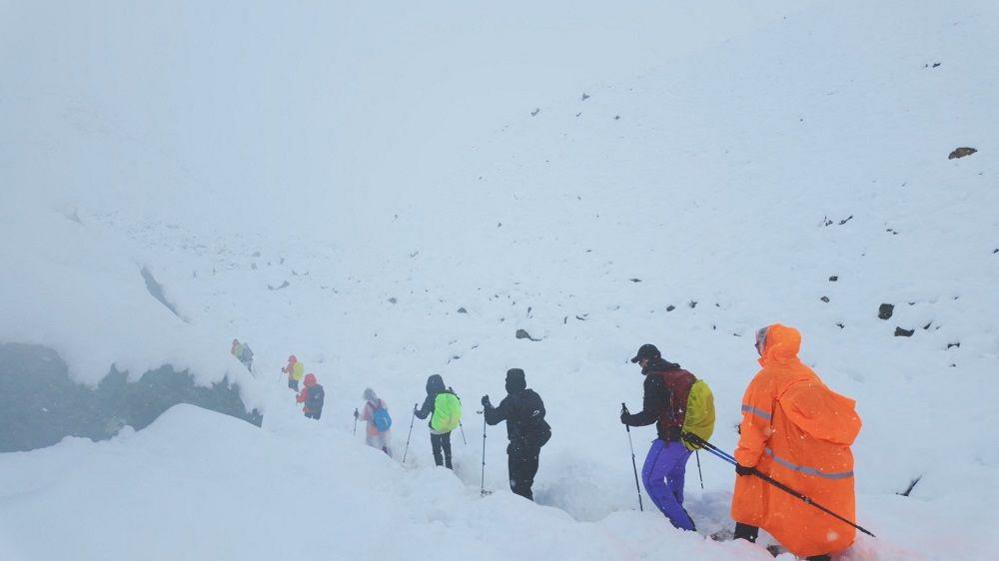 Backview of hikers in winter coats using hiking sticks to walk up a snowy slope. They're surrounded by snowy, mountainous terrain.