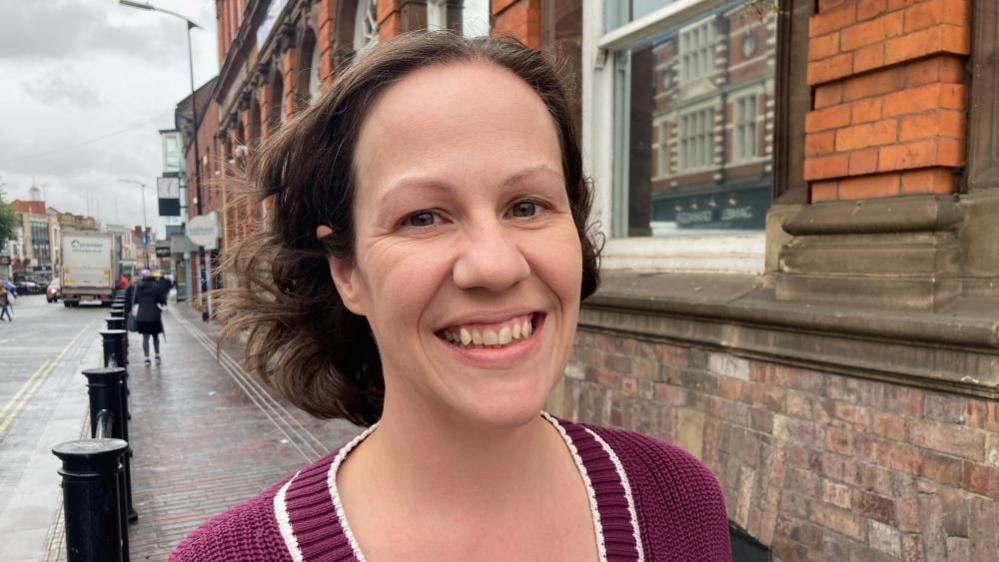 Lauren Ellis, who has long, dark hair and is wearing a purple top, is smiling at the camera. There is a brick building to her left and a rain-soaked shopping street to her right.