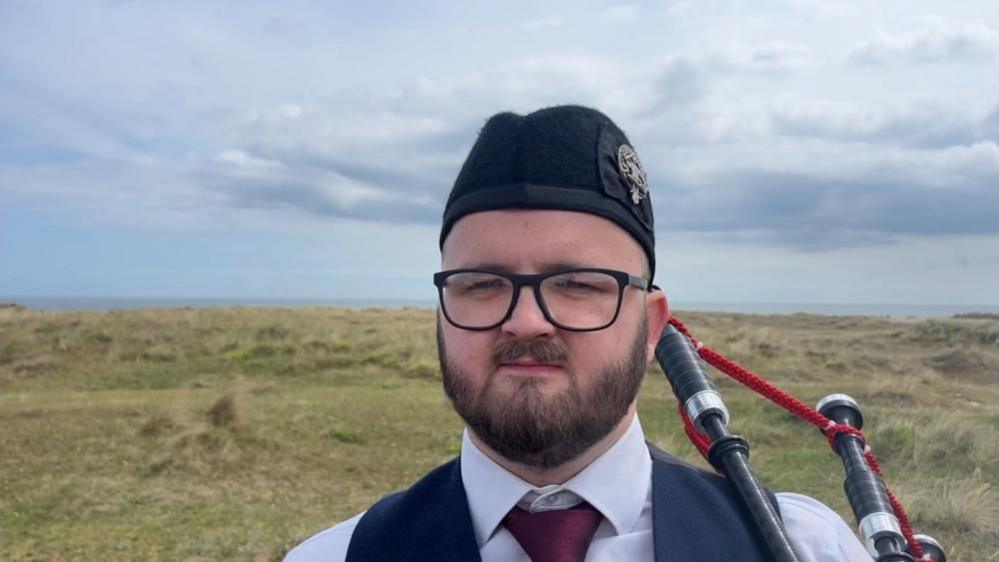 Ryan Fitzpatrick stands on the marram grass of the North Denes Beach in Great Yarmouth. He is wearing a black hat, white shirt, red tie, dark blue waistcoat. He is wearing black rimmed glasses, and sports a full beard and moustache. He is holding his bagpipes, with the pipes over his shoulder, tethered with red cord.
