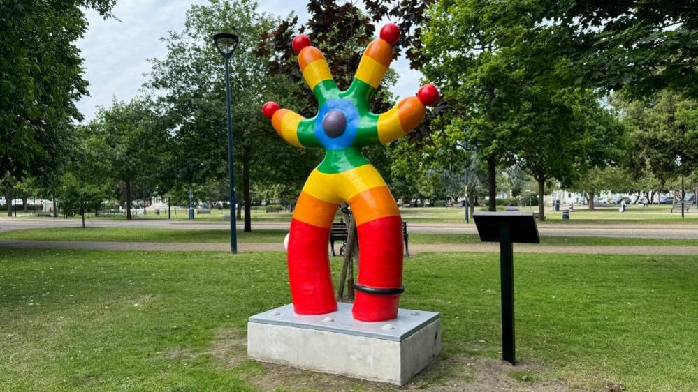 A four-armed, two legged sculpture painted in concentric bands (from outer to inner) red, orange, yellow, green, blue and purple. The design has a ball in the middle and on the tips of all four arms. 
