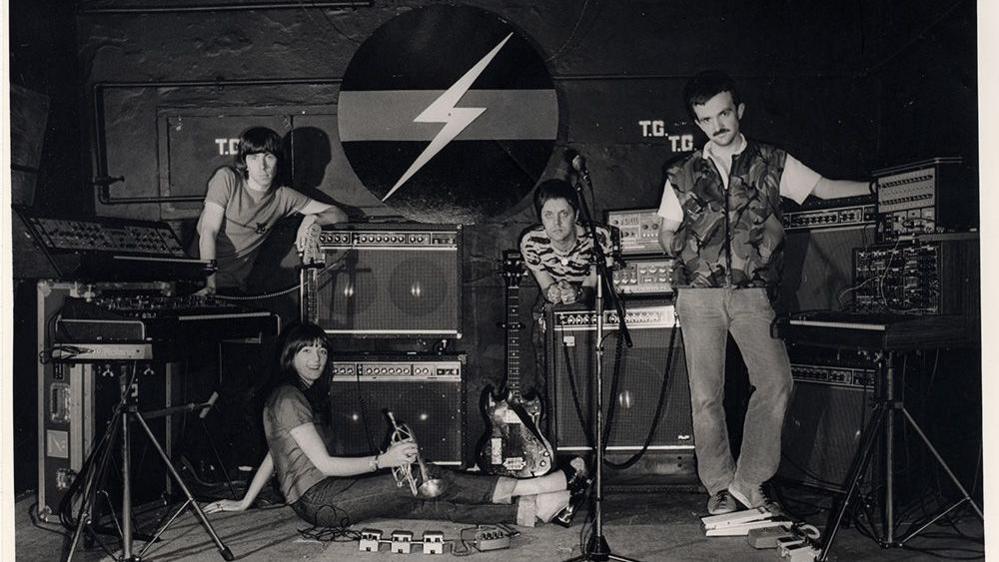 Black and white picture of four musicians. There are three men and a woman who is sitting. They are surrounded by instruments with a logo behind them depicting a flash. The woman is holding a cornet.