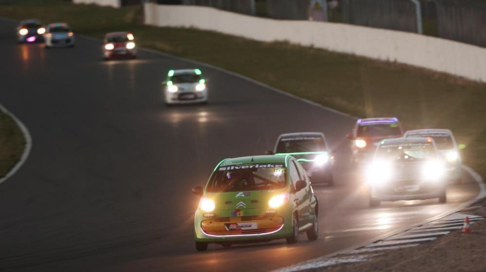Silverstone Circuit at night with Citroen C1 cars of various colours taking a banked corner with their headlights shining. The front car is green, with a Silverlink banner across the top of the windscreen. There is a group of four cars close behind the leader.