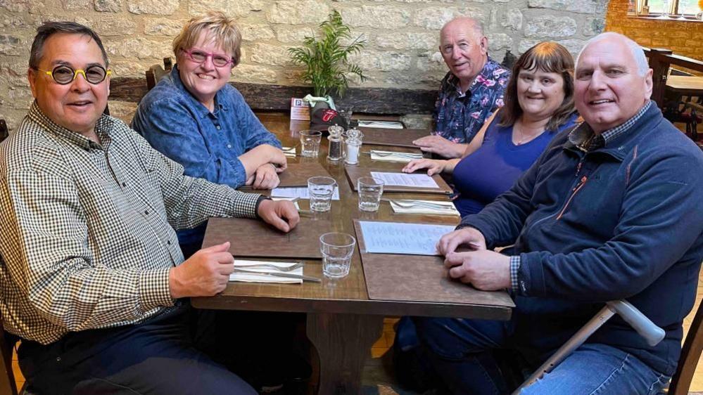 Five people, sitting round a table, in a pub. They have menus and glasses in front of them. They are all smiling and looking at the camera. There is a stone wall behind them. 