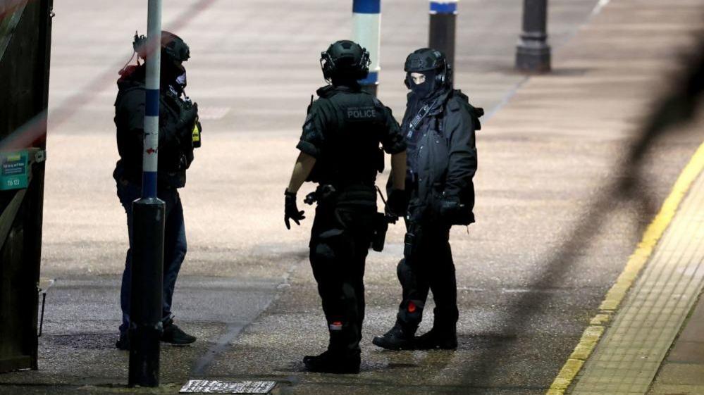 Three specially trained officers dressed in dark clothing, helmets and gloves stand on a train station platform