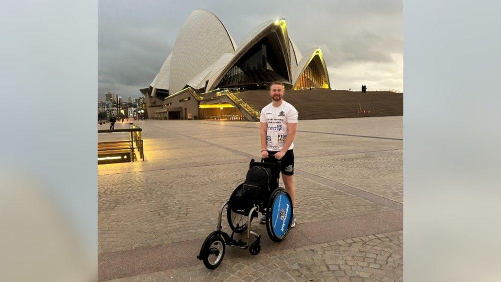 Billy Whiston is standing with his wheelchair in front of the Sydney Opera House, at the start of his journey.