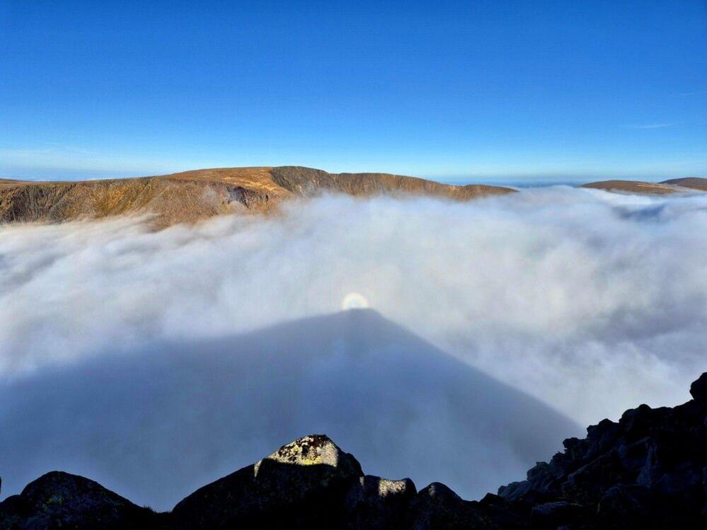 The view from one munro peak to another is obscured by low lying clouds. Above these clouds is a bright blue sky. There are halo-like rings of rainbow-coloured light in the centre of the photo. 