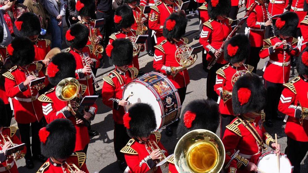 An aerial shot of the Coldstream Guards marching through Berwick. They are wearing full uniform with bearskin hats and bright red military jackets. All are playing musical instruments including wind instruments and drums.