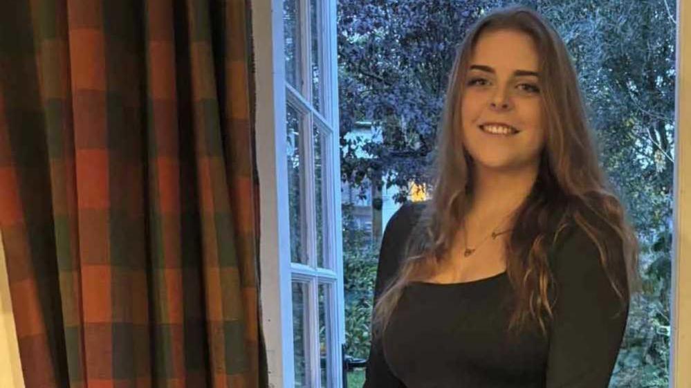 A woman with long brown hair and wearing a black long sleeved T-shirt and a necklace stood by a glass door with trees outside. There is a red and black checked curtain to her left.