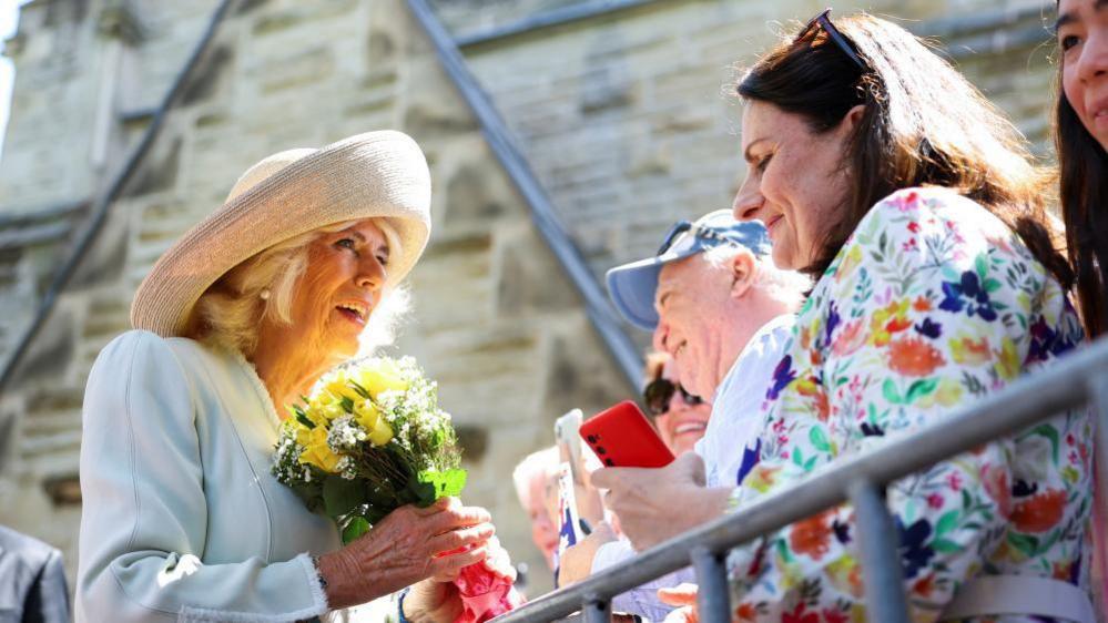 Queen Camilla is seen on the left of the image, facing towards the right. She is wearing a bright blue suit and a beige hat. She is holding a small bunch of yellow flowers. Camilla is speaking to someone in the crowd.