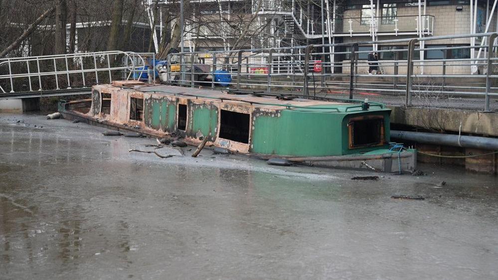 A red and green barge which has been badly damaged by fire sits on a frozen body of water beside a jetty.