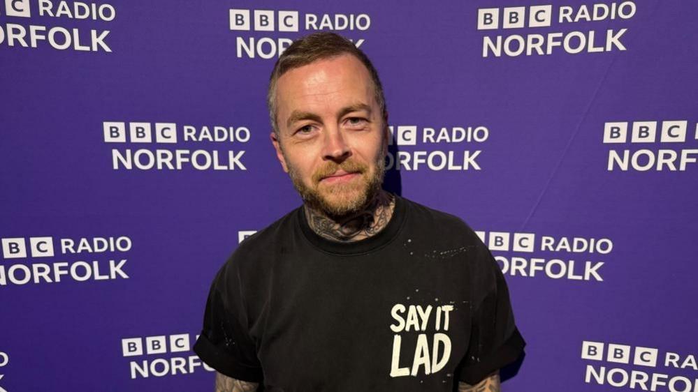 Jonny Human has short dark hair combed up on top and cropped short on the sides. He has a light brown full beard and moustache. He is wearing a Say It Lad branded tee shirt, and is standing in front of a BBC Radio Norfolk branded wall.