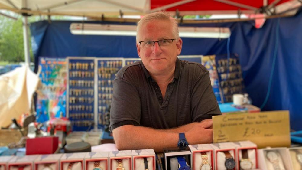 Les Brennan with short white hair and glasses, stands in his key-cutting stall