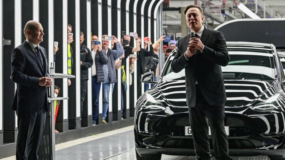 Elon Musk on the right stands holding a microphone in front of a Tesla car at his factory near Berlin in 2022