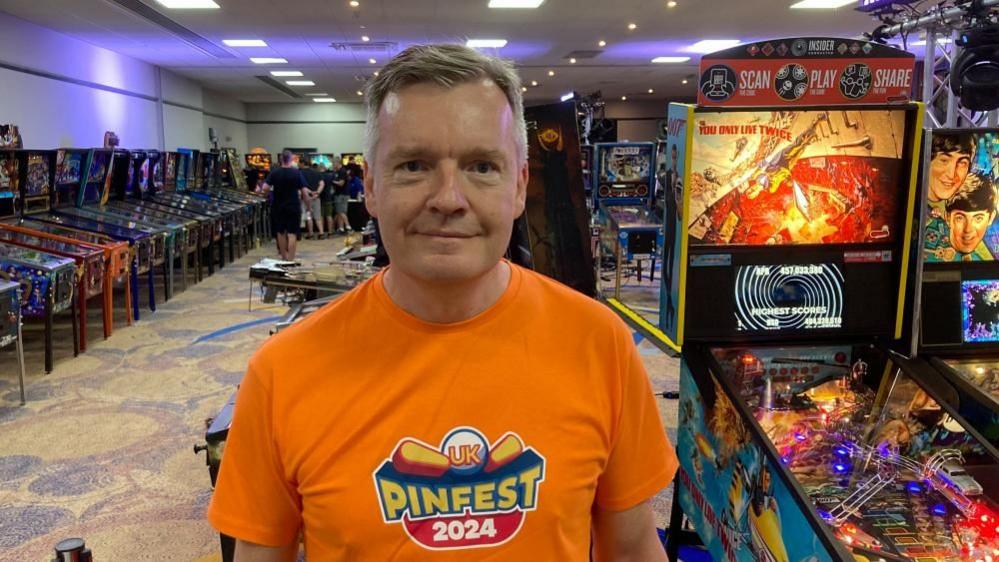 Philip Murphy with short dark hair wearing an orange Pinfest tee shirt with pinball machines behind