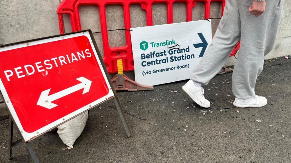 A sign saying Belfast Grand Central Station in navy letters on the ground beside an orange roadworks barrier and a red pedestrians sign.