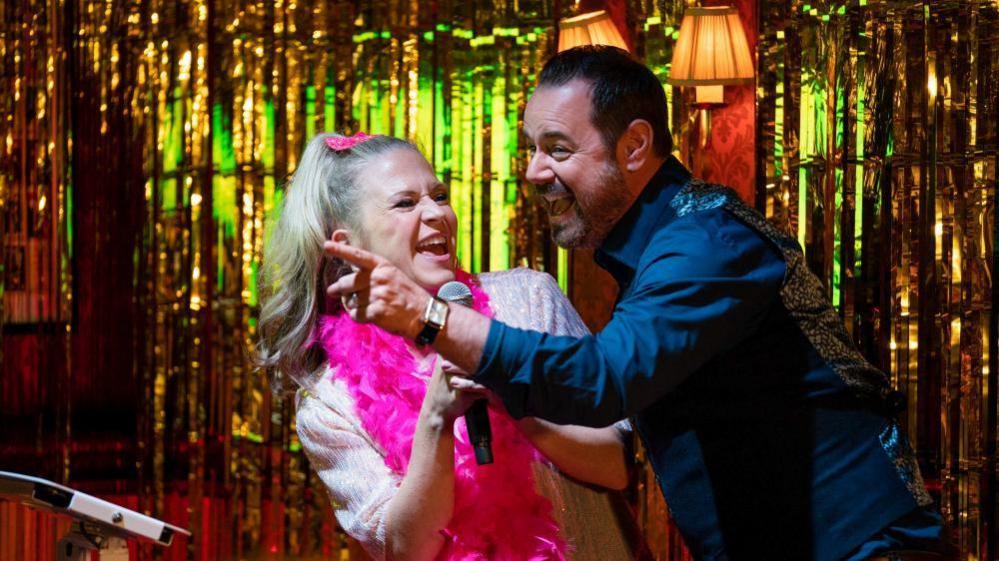 A blonde woman smiles broadly while wearing a pink feather boa and holding a karaoke microphone. She is stood on a glittery stage with a brown hair man who is also smiling. 