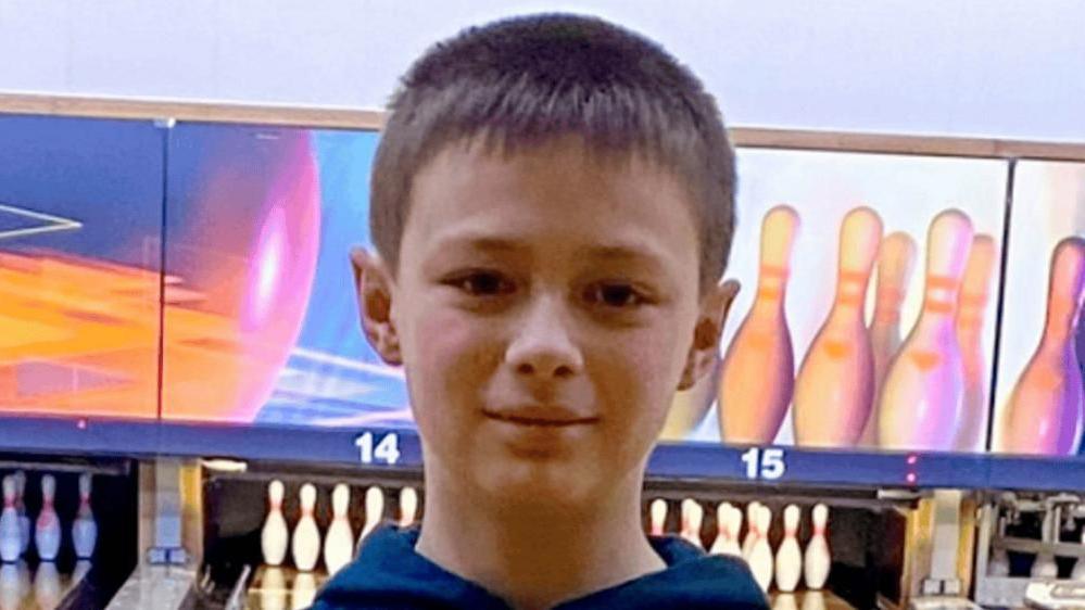 A headshot of a 12-year-old boy, who is smiling into the camera and has short brown hair. He is standing in front of a bowling lane and the pins can be seen behind him. 