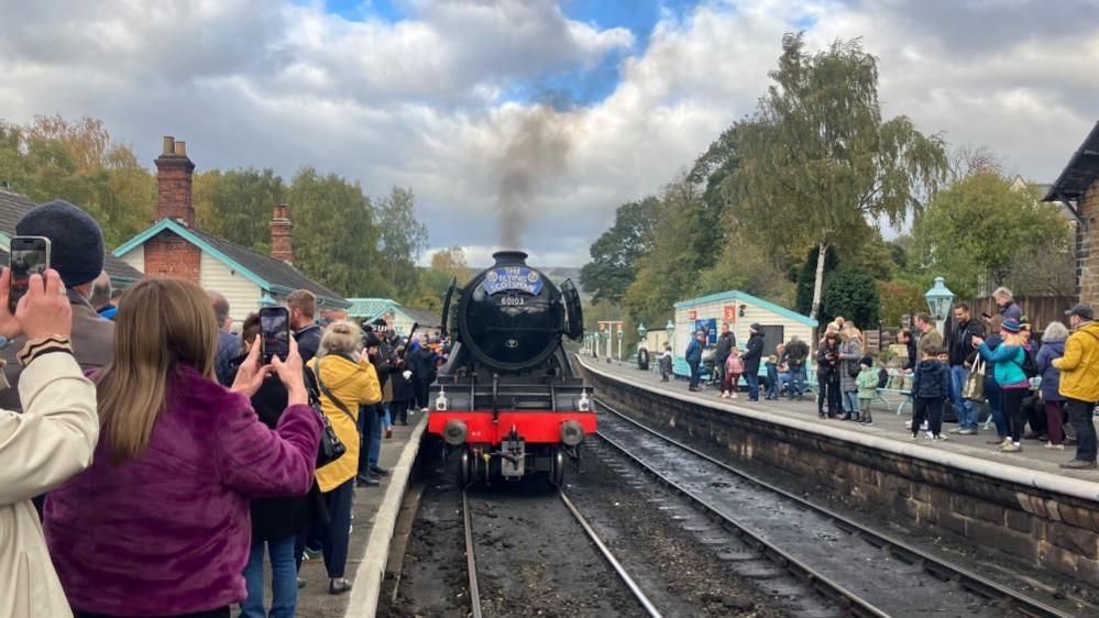 The front a a black stream train can be seen on railway tracks with crowds of people gathered on either side holding their phones and cameras up to take pictures. The engine appears to be 'in steam' with black smoke coming from the chimney. The blue nameplate on the engine reads 'The Flying Scotsman' and the number 60103 can be seen in gold print.