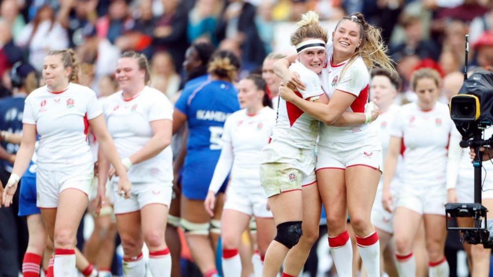 England's Megan Jones and Jess Breach celebrate after qualifying for the final.. They are wearing the white kit and hugging with other team members behind them and a camera to the right. Crowds are in the distance. 