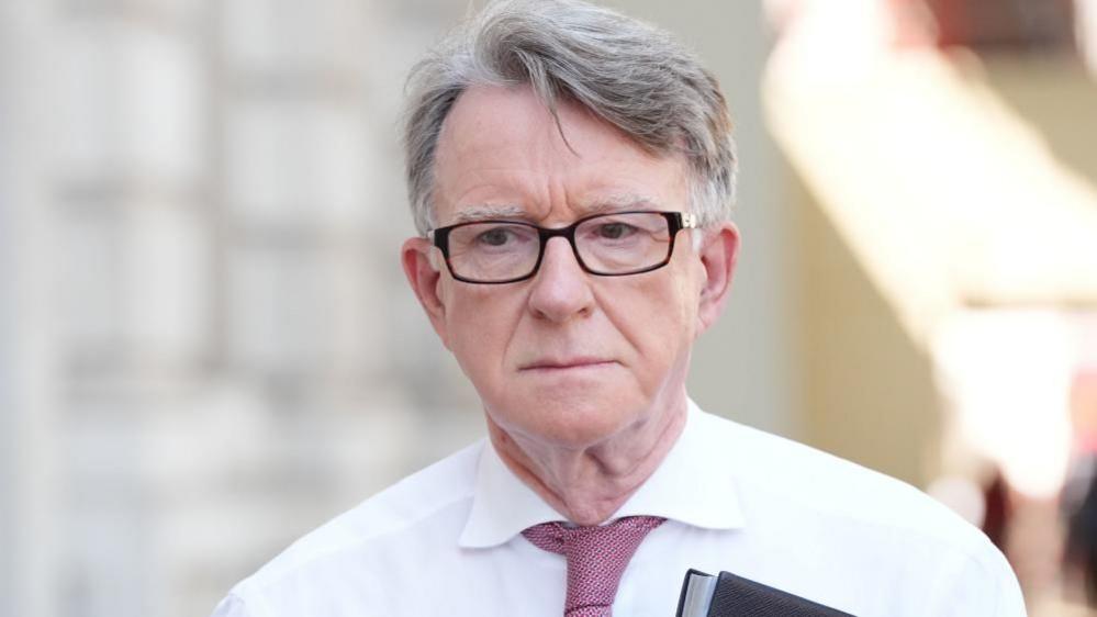 Lord Mandelson arriving at the Cabinet Office in central London. He is wearing a white short, pink tie, dark rimmed glasses and is holding a file with a black cover.