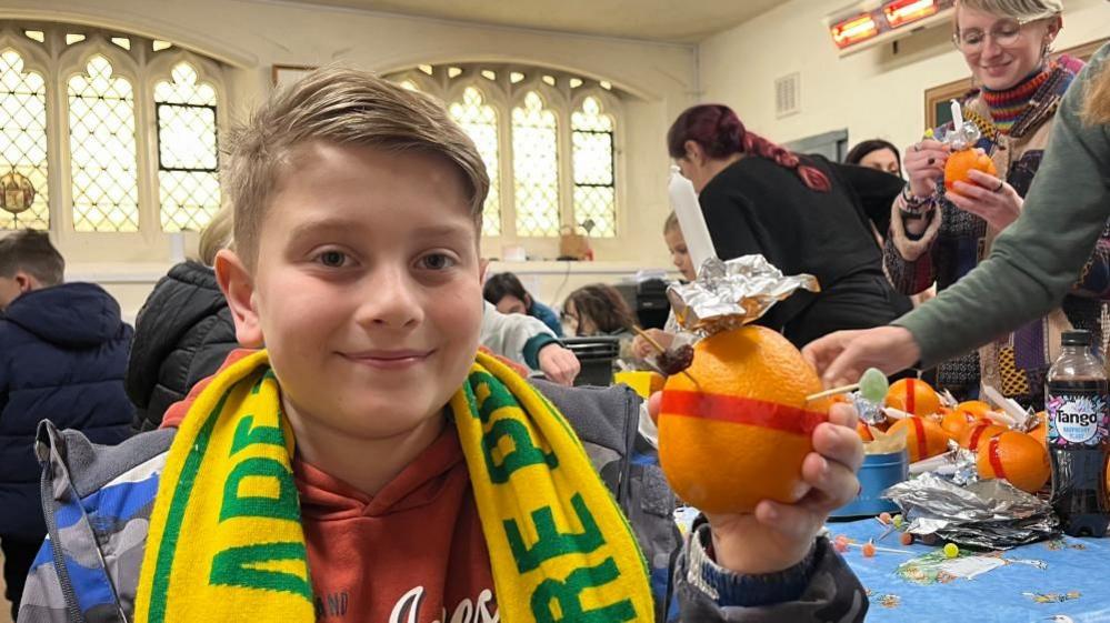 Jonah holds one of many Christingles he made during a workshop at Great Yarmouth Minster on Monday. Jonah, has blonde hair and is wearing a yellow and green Norwich City Football Club scarf. 